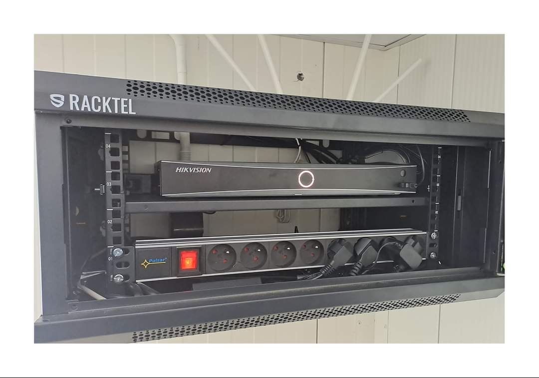 Zamknięta szafa Racktel z zamontowanym rejestratorem Hikvision i listwą zasilającą Pulsar. Widoczne kable i elementy instalacji. Całość zamontowana na białej ścianie.