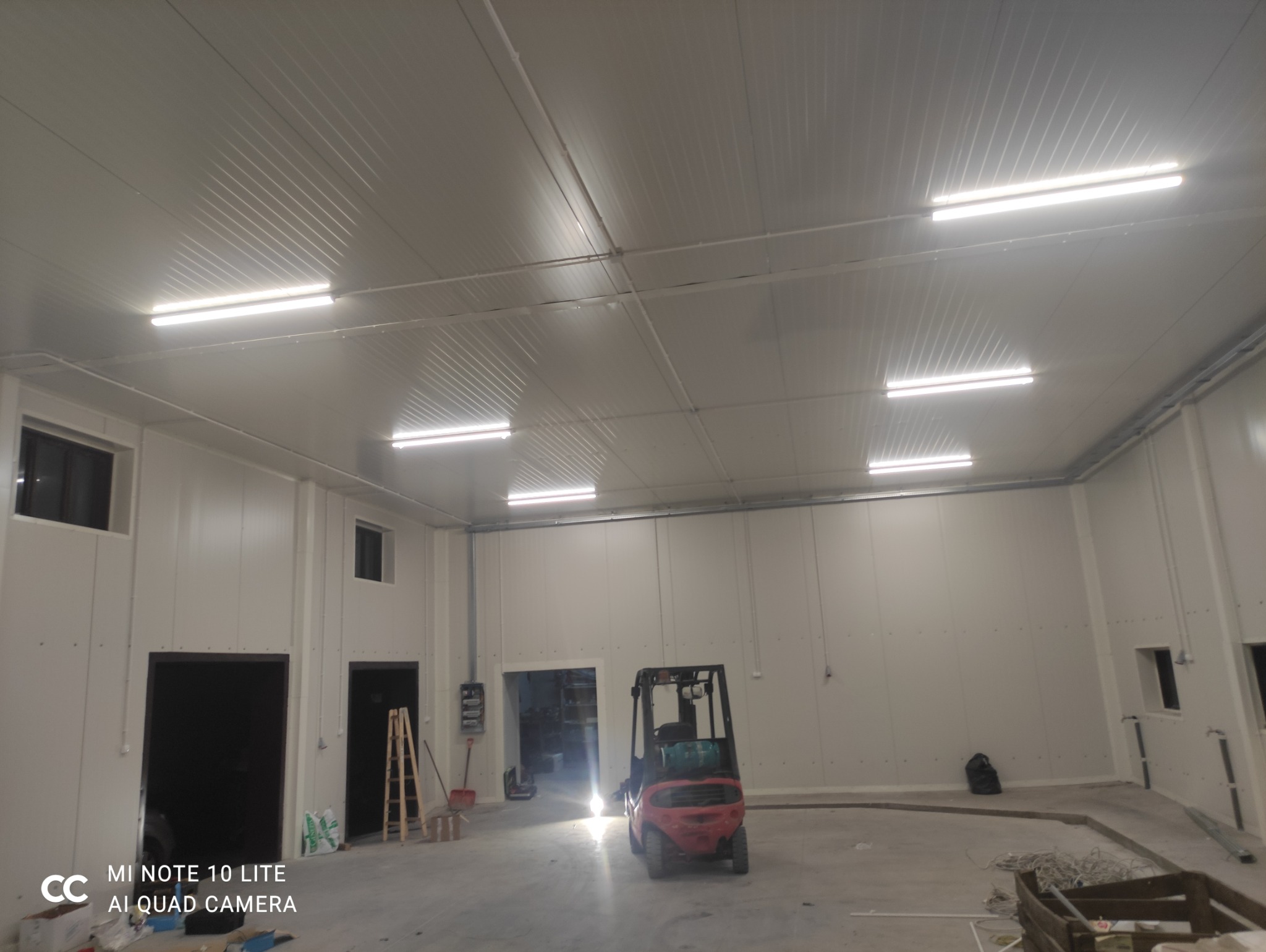 Wnętrze hali z zamontowanym oświetleniem liniowym. Widoczny wózek widłowy, drabina i okna. Białe ściany i sufit z widocznymi panelami.