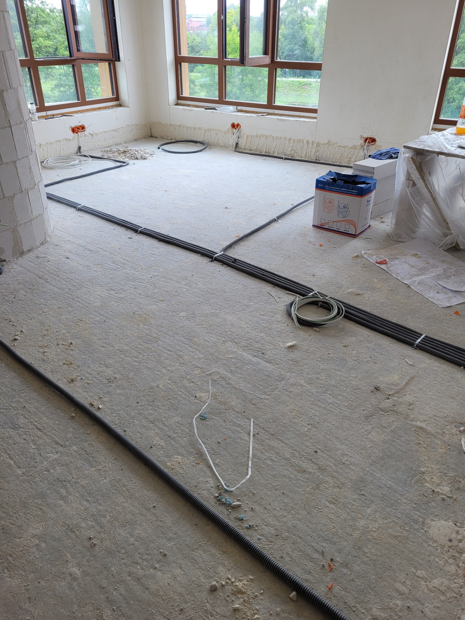 Wnętrze w trakcie prac elektrycznych: kable w peszlach ułożone na podłodze, puszki instalacyjne w ścianach, okna z widokiem na zieleń. Etap przed wylewką.