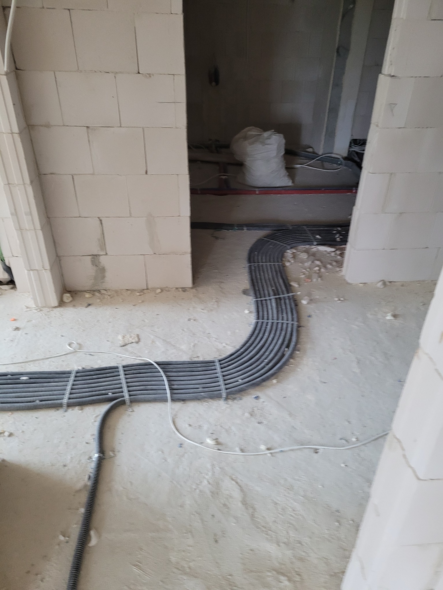 Szare rury instalacji elektrycznej ułożone równolegle na podłodze w surowym wnętrzu budynku z białych bloczków, widoczne kable i fragmenty ścian.