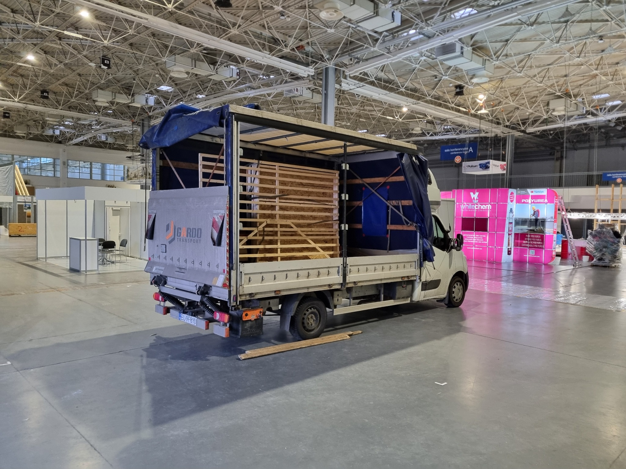 Ciężarówka transportowa z otwartą przestrzenią ładunkową wypełnioną drewnianymi elementami, zaparkowana w hali wystawienniczej z widocznymi stoiskami w tle.