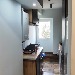 DOM-MAJSTER Michał Siemieniuk - Wnętrze kuchni po remoncie z brązowymi płytkami podłogowymi, białymi ścianami, zabudową kuchenną z drewnianymi frontami i czarnymi akcentami oraz oświetleniem punktowym.