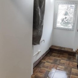 DOM-MAJSTER Michał Siemieniuk - Pokój w trakcie prac: świeżo pomalowane ściany na biało, zabezpieczony grzejnik, brązowa, błyszcząca podłoga z płytek imitujących drewno ułożonych w kwadraty, widoczne kable elektryczne wystające...