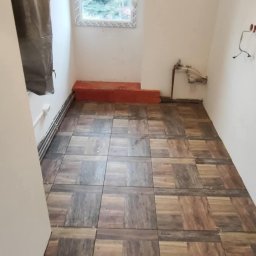 DOM-MAJSTER Michał Siemieniuk - Ułożona podłoga z płytek imitujących drewno w pomieszczeniu w Lubaniu, widoczne niedociągnięcia w spoinach i przygotowane do położenia płytki przy ścianie.