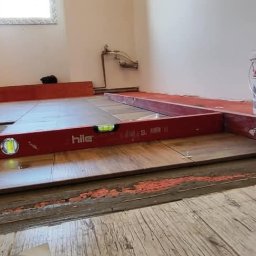 DOM-MAJSTER Michał Siemieniuk - Układanie podłogi z płytek imitujących drewno, poziomowane czerwoną poziomicą Hile, widoczne krzyżyki dystansowe, wiadro z klejem Fast A i ślady zaprawy na podłożu.