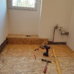 DOM-MAJSTER Michał Siemieniuk - Surowy stan łazienki w trakcie remontu: widoczna podłoga z płyty OSB, narzędzia (wiertarka, poziomica, młotek), rury instalacyjne przy ścianie, okno w tle.