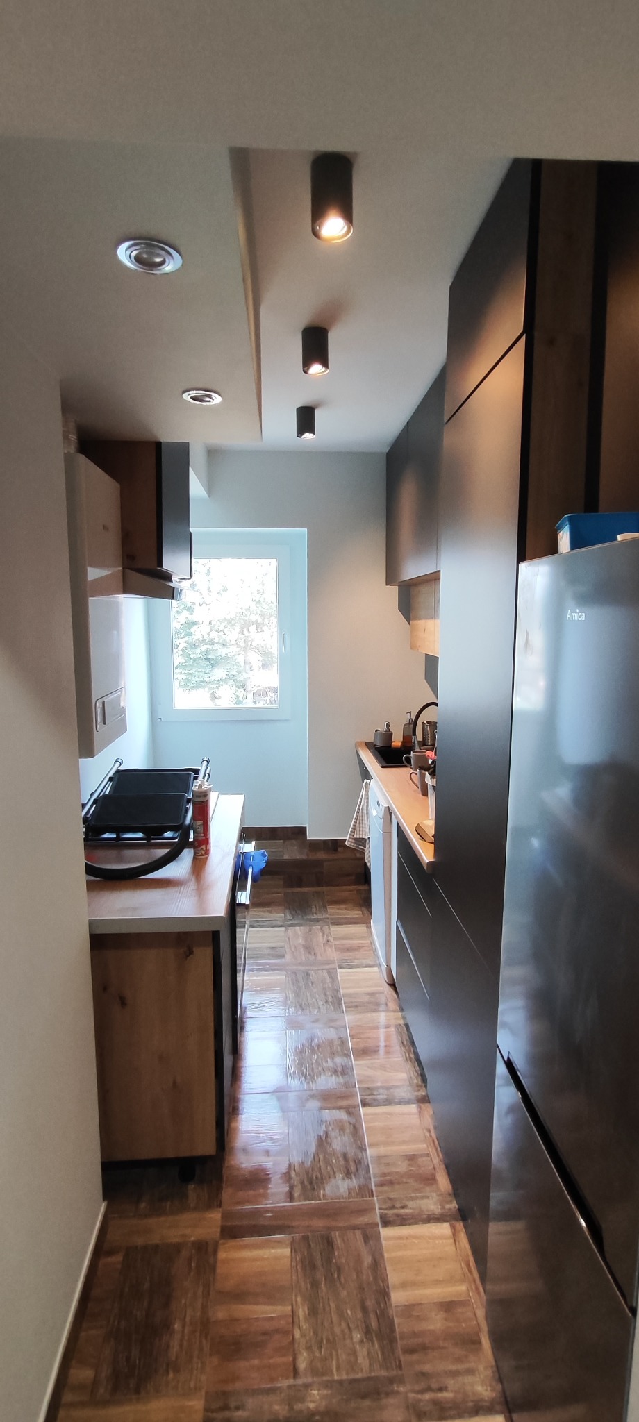 Wąska kuchnia z drewnianą podłogą imitującą panele, ciemnymi szafkami i oświetleniem punktowym na suficie.