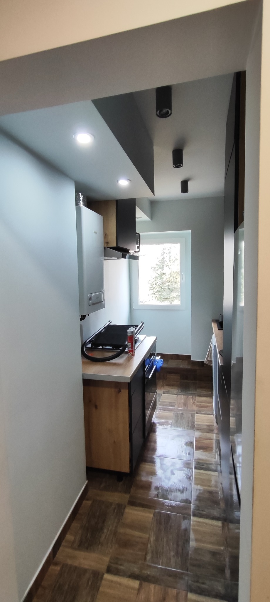 Wnętrze kuchni po remoncie z brązowymi płytkami podłogowymi, białymi ścianami, zabudową kuchenną z drewnianymi frontami i czarnymi akcentami oraz oświetleniem punktowym.