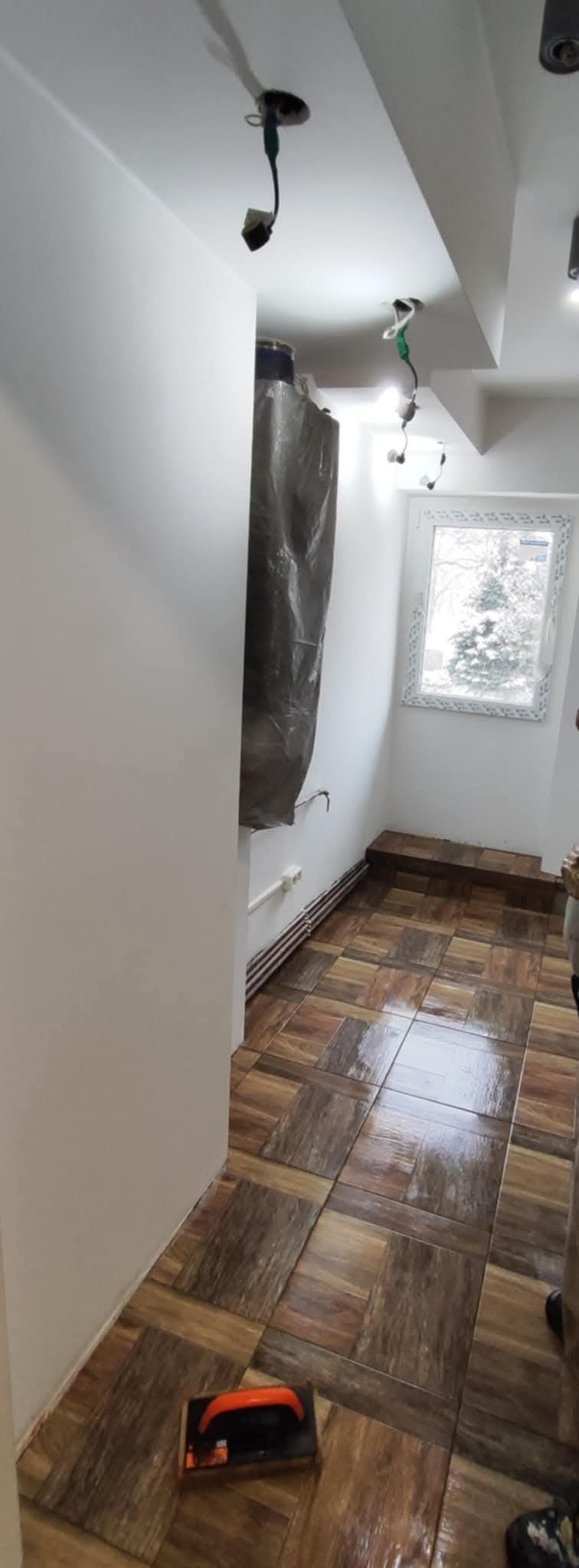 Pokój w trakcie prac: świeżo pomalowane ściany na biało, zabezpieczony grzejnik, brązowa, błyszcząca podłoga z płytek imitujących drewno ułożonych w kwadraty, widoczne kable elektryczne wystające...