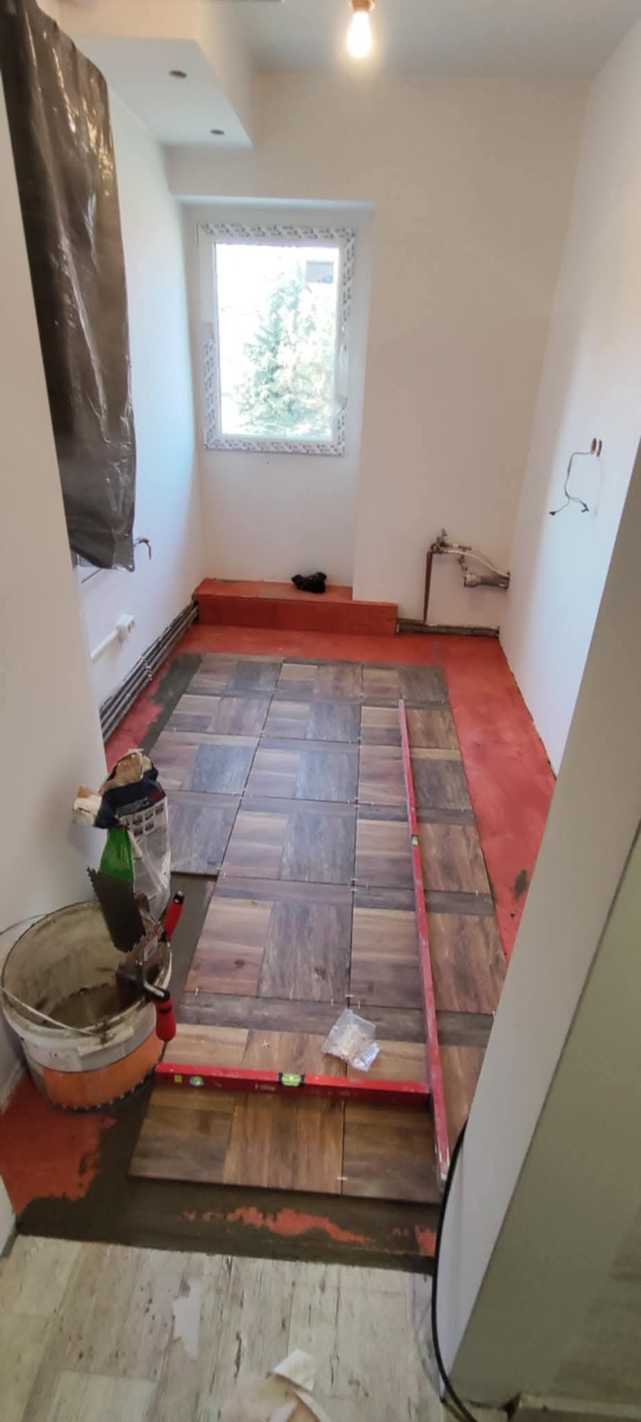 Układanie brązowych płytek podłogowych w łazience, widoczny poziomica, wiadro z zaprawą i paczka krzyżyków dystansowych.