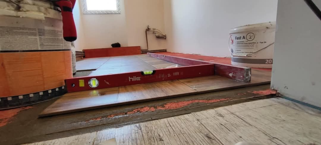 Układanie podłogi z płytek imitujących drewno, poziomowane czerwoną poziomicą Hile, widoczne krzyżyki dystansowe, wiadro z klejem Fast A i ślady zaprawy na podłożu.