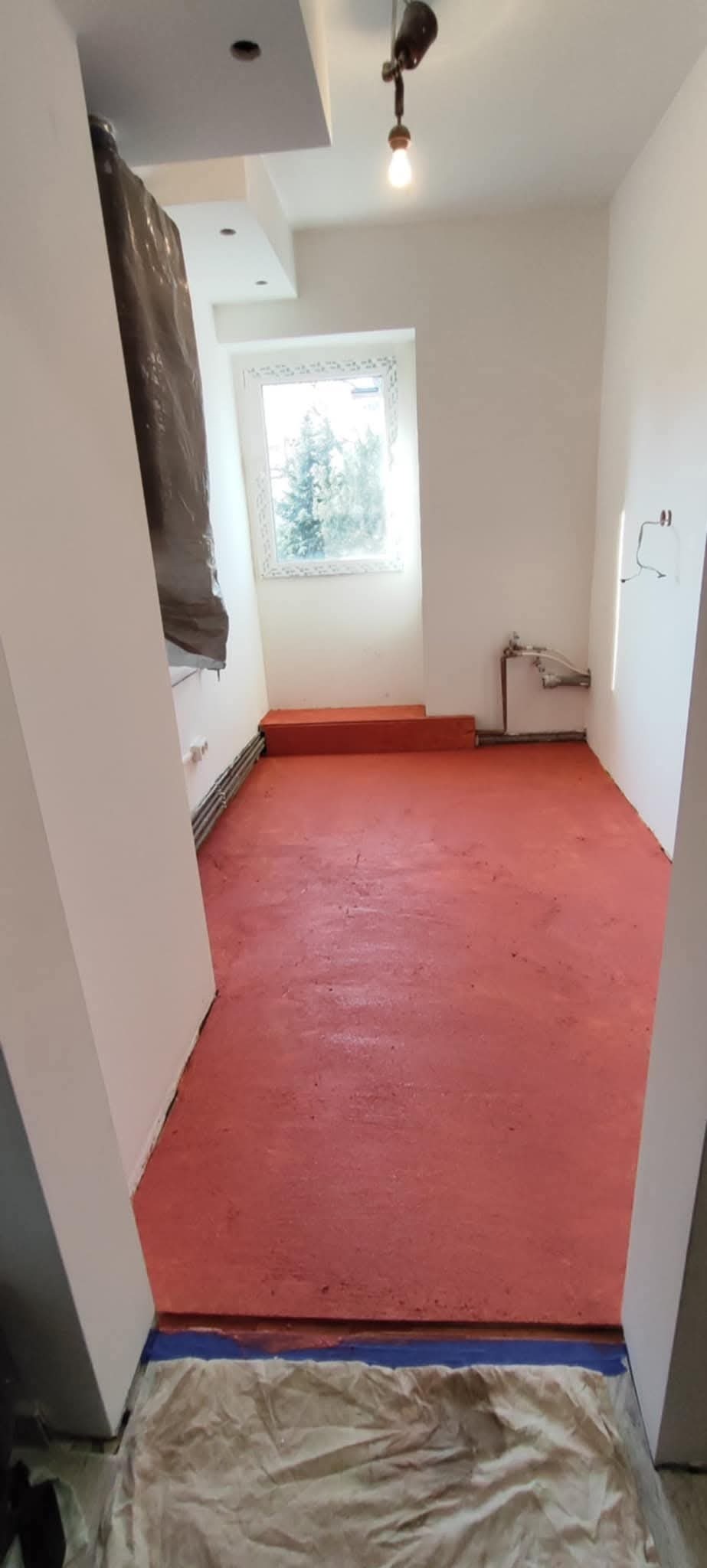 Pokój w trakcie prac przygotowawczych do malowania podłogi na czerwono, zabezpieczone ściany i podłoga folią malarską, widoczne grzejniki i instalacja elektryczna.