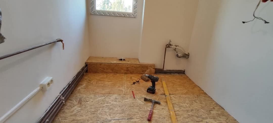Surowy stan łazienki w trakcie remontu: widoczna podłoga z płyty OSB, narzędzia (wiertarka, poziomica, młotek), rury instalacyjne przy ścianie, okno w tle.