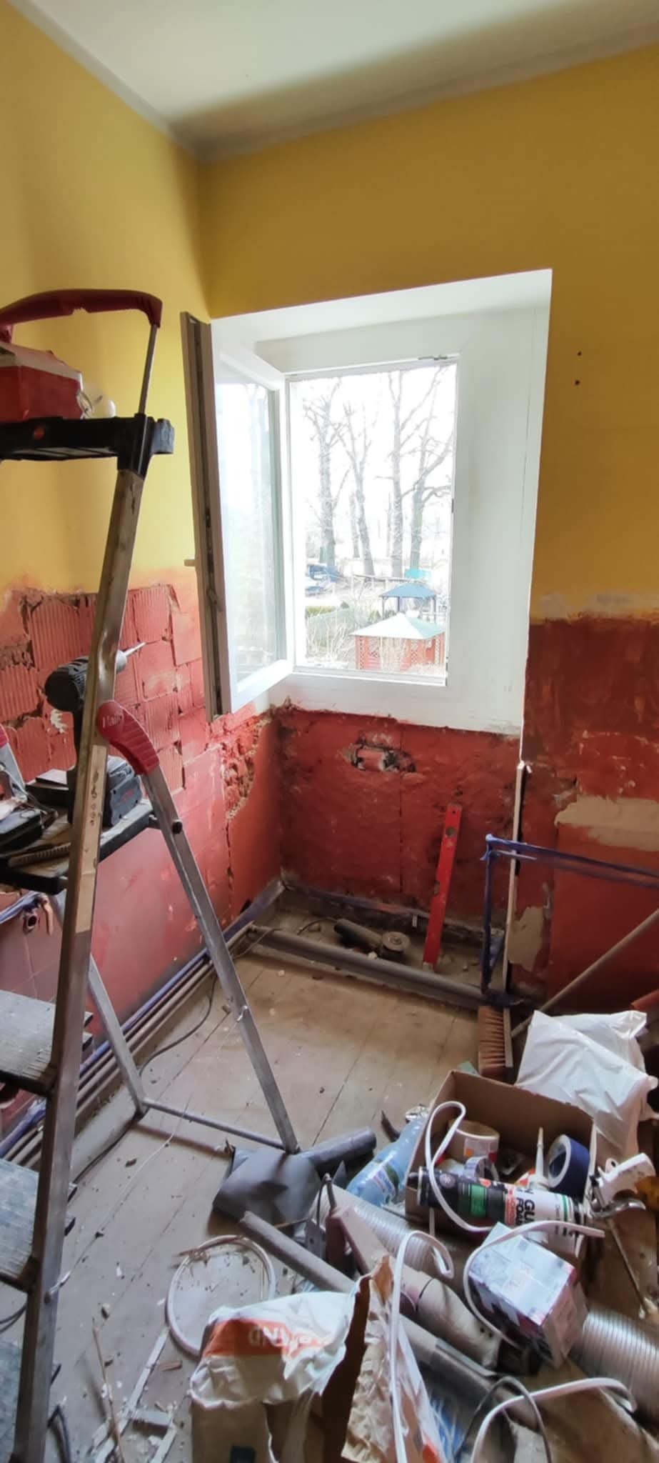 Pokój w trakcie remontu, widoczne odsłonięte cegły wokół okna, drabina aluminiowa i narzędzia w nieładzie, żółte ściany częściowo przygotowane do dalszych prac.