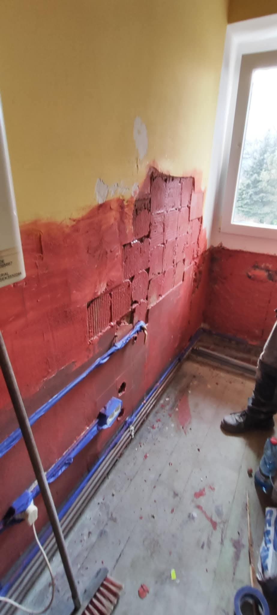 Przygotowanie ścian do położenia płytek w łazience: ściana z resztkami starej farby, nałożony czerwony grunt, widoczne kable elektryczne zabezpieczone niebieską taśmą malarską.