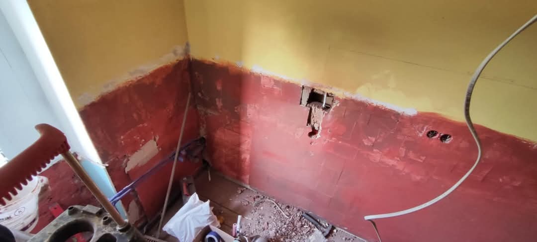 Fragment ściany w trakcie przygotowania do remontu, z widocznymi śladami starej farby, otworami na gniazdka elektryczne i wystającą instalacją elektryczną. Na podłodze widoczne gruz i narzędzia.