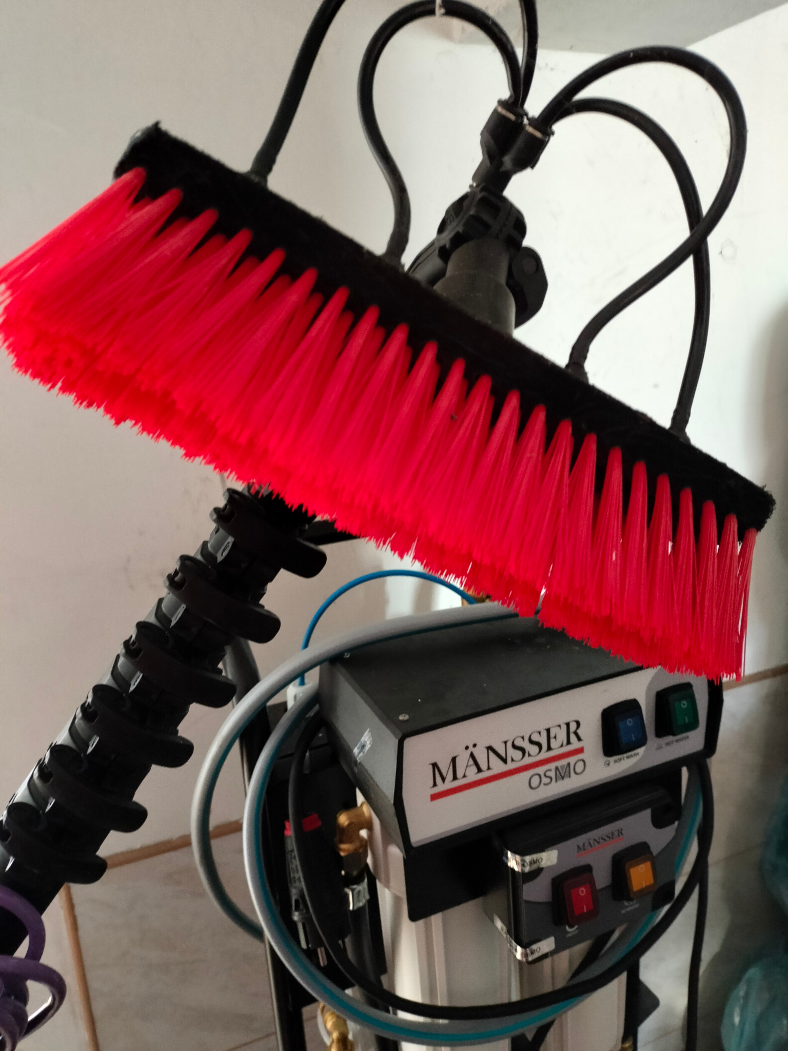 Szczotka z jaskrawoczerwonym włosiem do mycia okien, podłączona do urządzenia filtrującego wodę marki MÄNSSER OSMO, z widocznymi przewodami i przełącznikami.