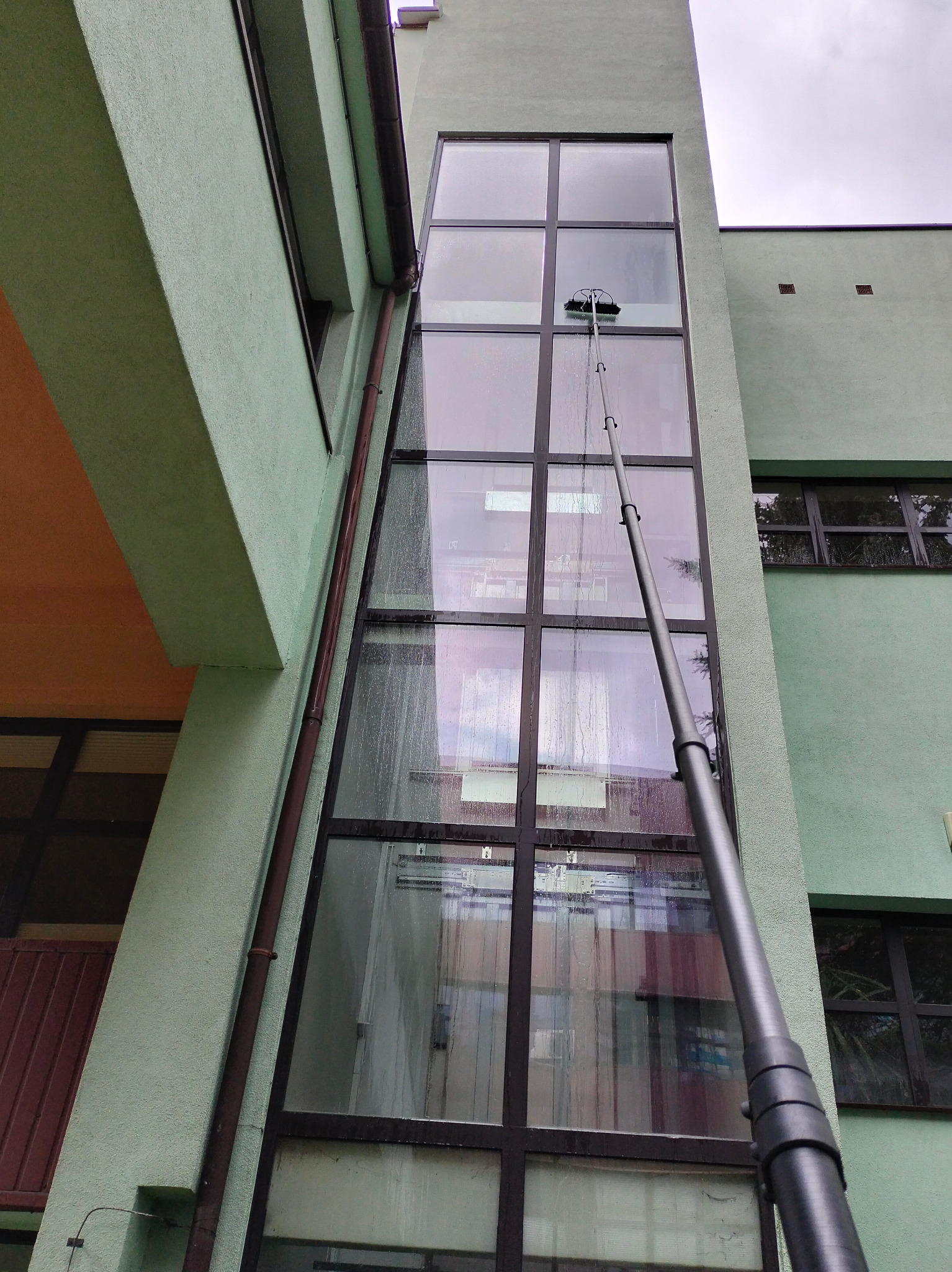 Mycie okien na wysokości budynku z zieloną elewacją za pomocą teleskopowej lancy z szczotką. Widoczne smugi wody na oknach, szara pogoda.