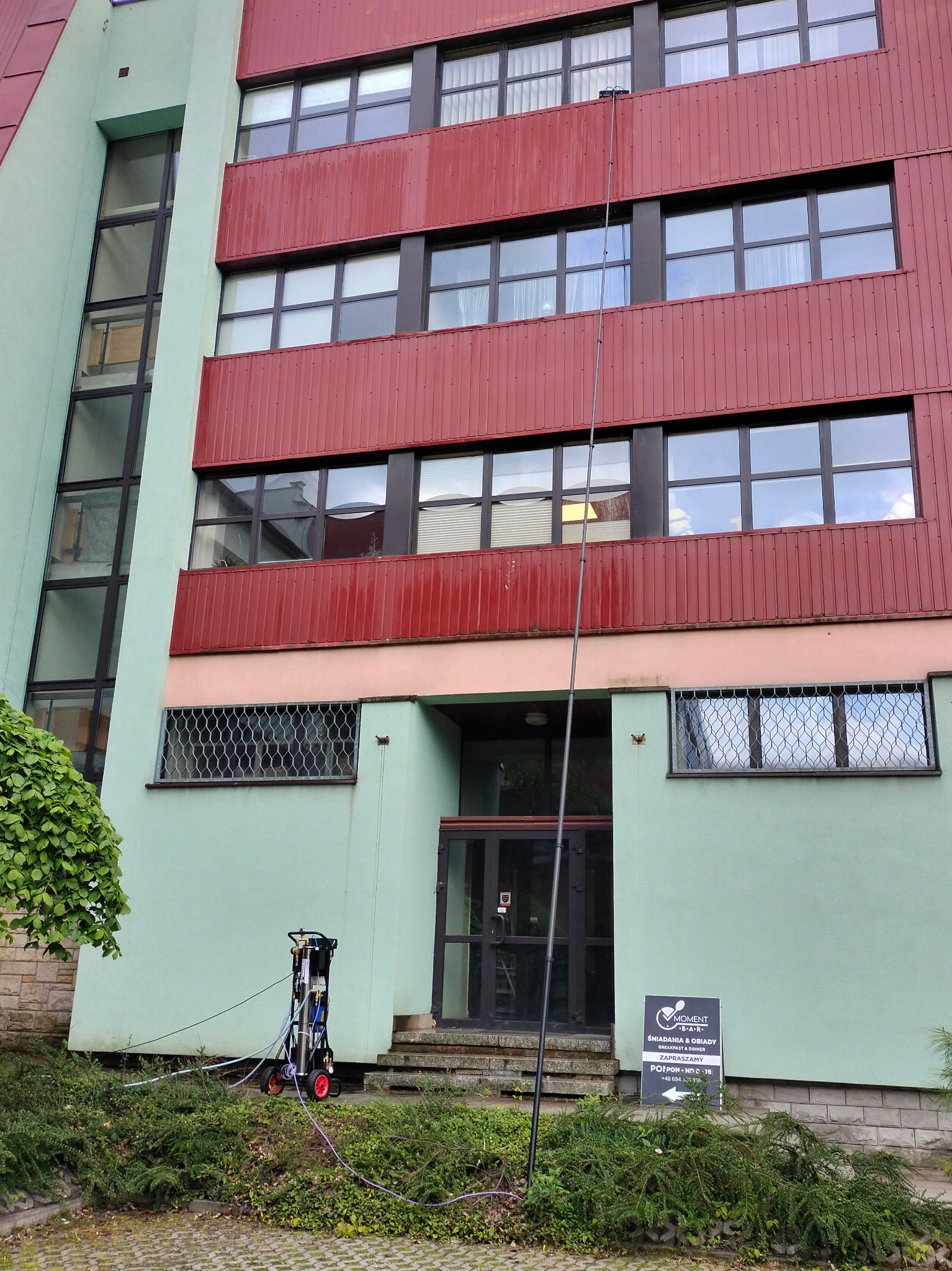 Budynek z oknami mytymi lancą teleskopową. Na dole wózek z urządzeniem do mycia. Widoczny szyld restauracji Moment.