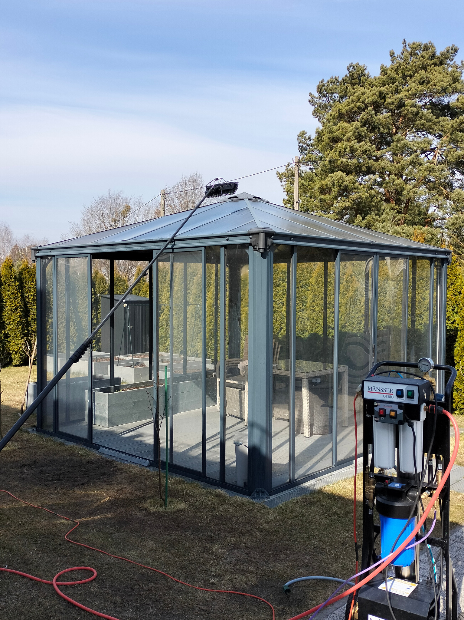 Mycie szklanej altany ogrodowej z użyciem specjalistycznego sprzętu. Widoczna lanca teleskopowa i urządzenie filtrujące wodę.