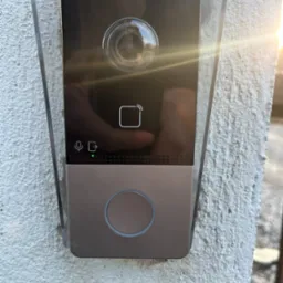 Zamontowany wideodomofon Hikvision w obudowie ochronnej, przymocowany do szarej, chropowatej ściany, z widocznym odbiciem w obiektywie kamery i świecącą zieloną diodą.