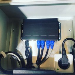 Cybite - Rozdzielnica z zamontowanym routerem, z podłączonymi kablami Ethernet z niebieskimi końcówkami oraz przewodem zasilającym, umieszczona w szarej skrzynce w pomieszczeniu z ceglanym murem.