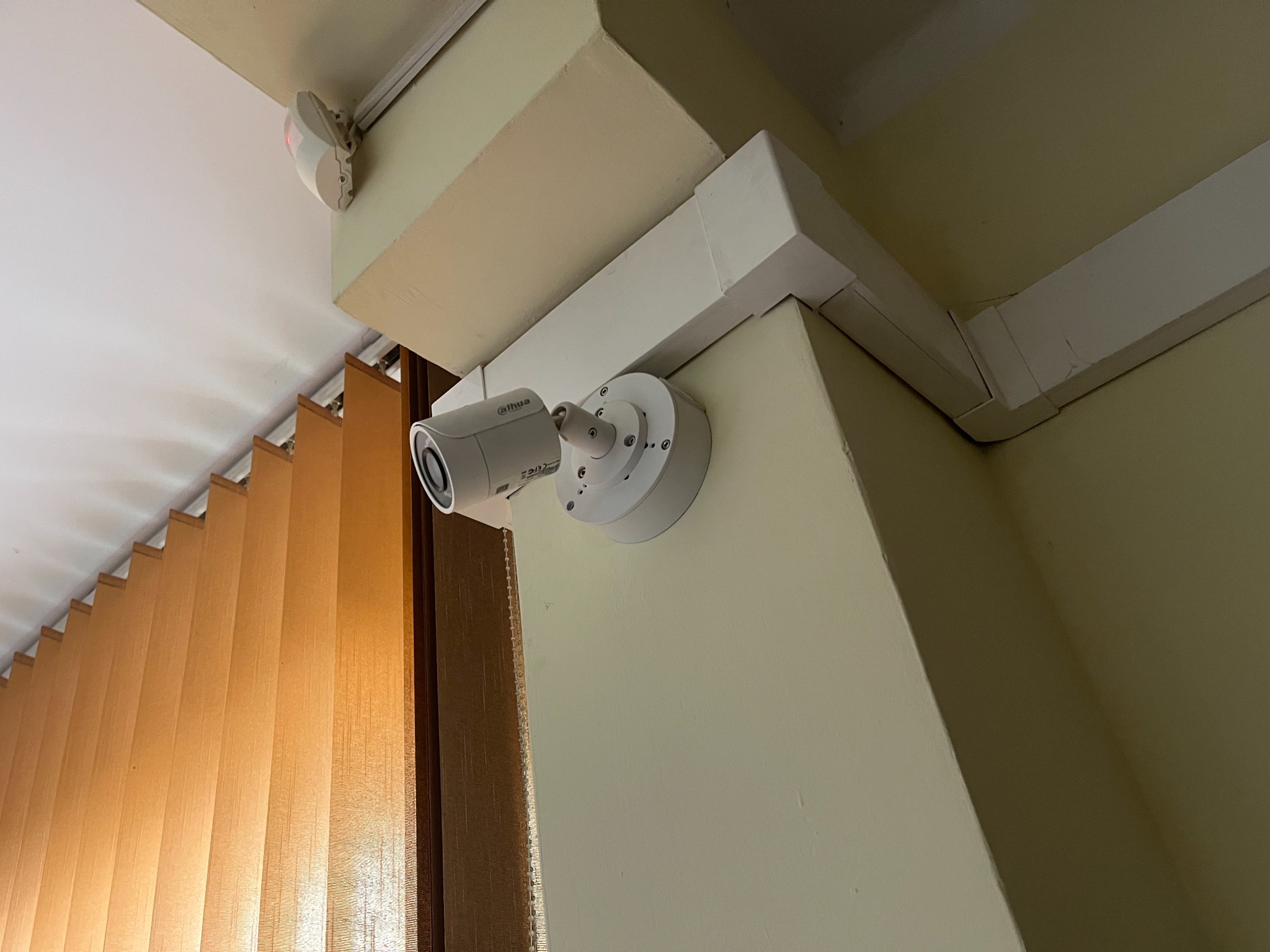 Kamera monitoringu Dahua zamontowana na ścianie w rogu pomieszczenia, widoczne brązowe żaluzje i czujnik ruchu pod sufitem.