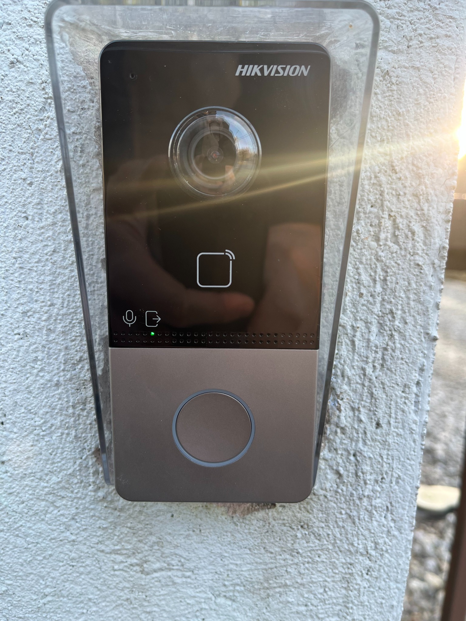 Zamontowany wideodomofon Hikvision w obudowie ochronnej, przymocowany do szarej, chropowatej ściany, z widocznym odbiciem w obiektywie kamery i świecącą zieloną diodą.