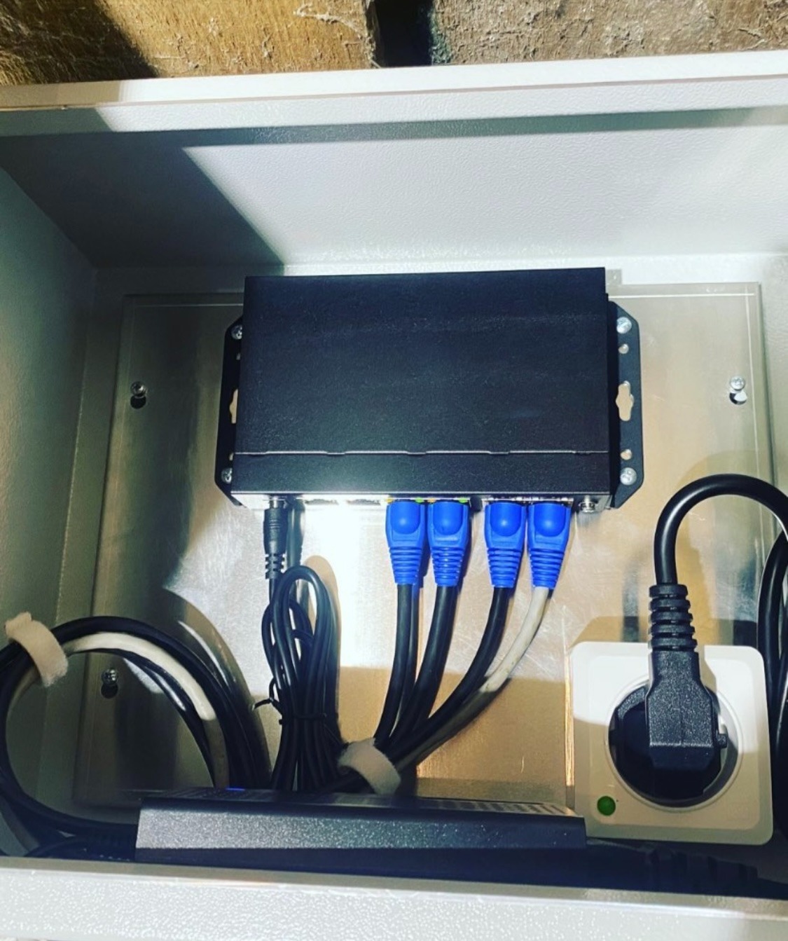 Rozdzielnica z zamontowanym routerem, z podłączonymi kablami Ethernet z niebieskimi końcówkami oraz przewodem zasilającym, umieszczona w szarej skrzynce w pomieszczeniu z ceglanym murem.