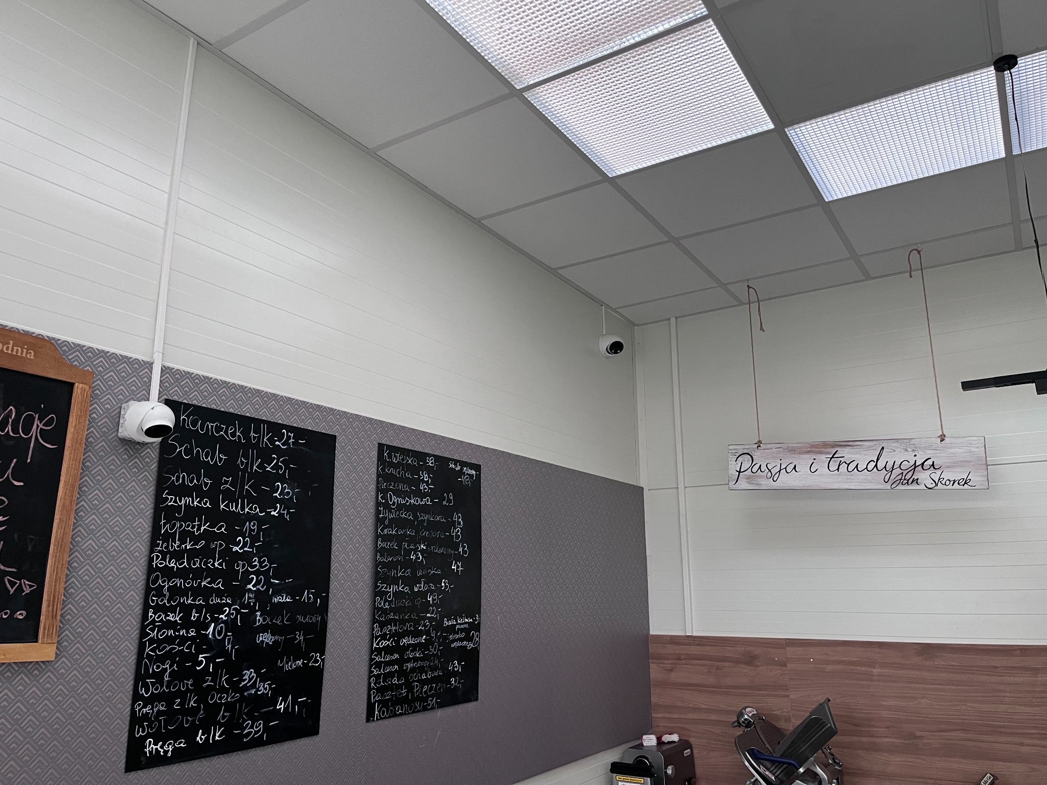 Kamera monitoringu zamontowana w sklepie mięsnym, widok z góry na tablice z cenami, napis 'Pasja i tradycja' oraz krajalnicę do wędlin.