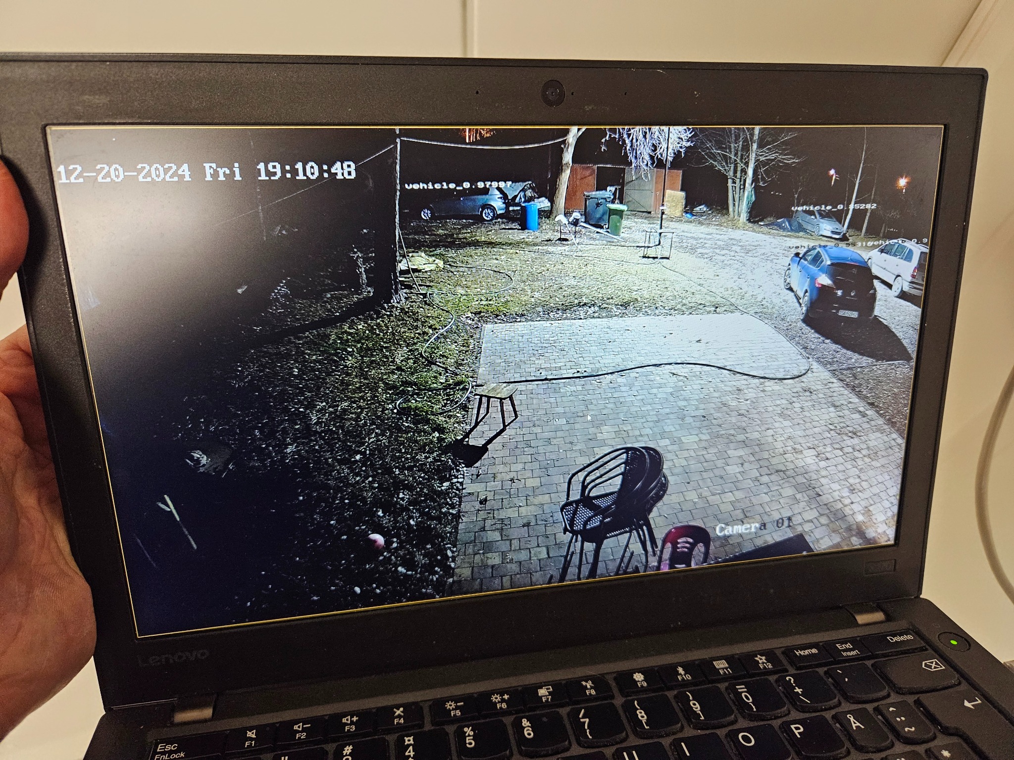 Ekran laptopa Lenovo wyświetlający obraz z kamery monitoringu posesji w nocy, z widocznymi samochodami, elementami wyposażenia ogrodu i datą 20 grudnia 2024.