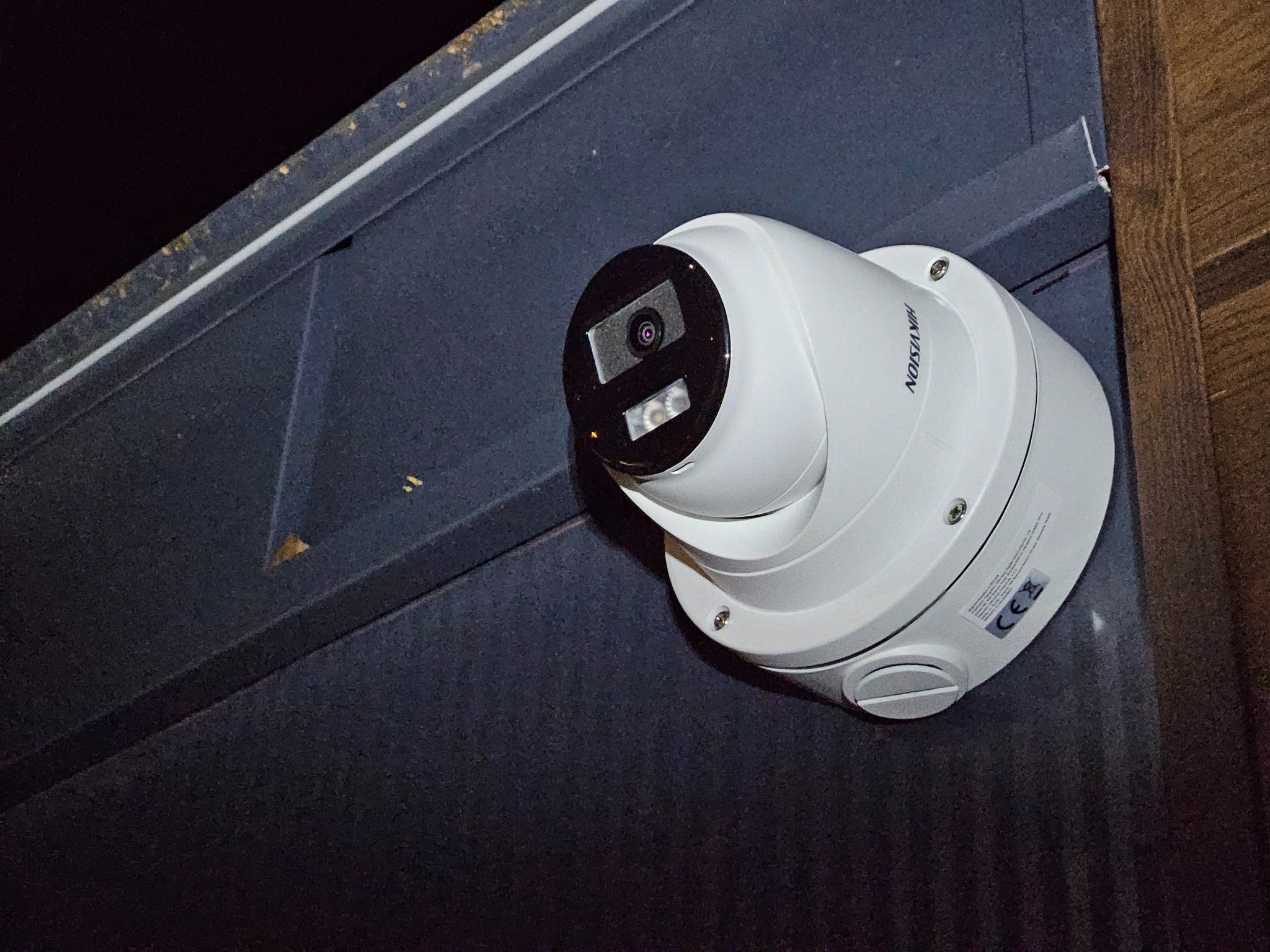Biała kamera kopułkowa Hikvision zamontowana na ciemnoszarej, metalowej powierzchni pod kątem, widoczne śruby mocujące i naklejka z certyfikatem CE.