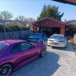 Trzy sportowe auta (fioletowy Nissan Skyline R34, niebieski Toyota MR2, srebrna Toyota Supra) zaparkowane przed garażem w słoneczny dzień, widok z boku.