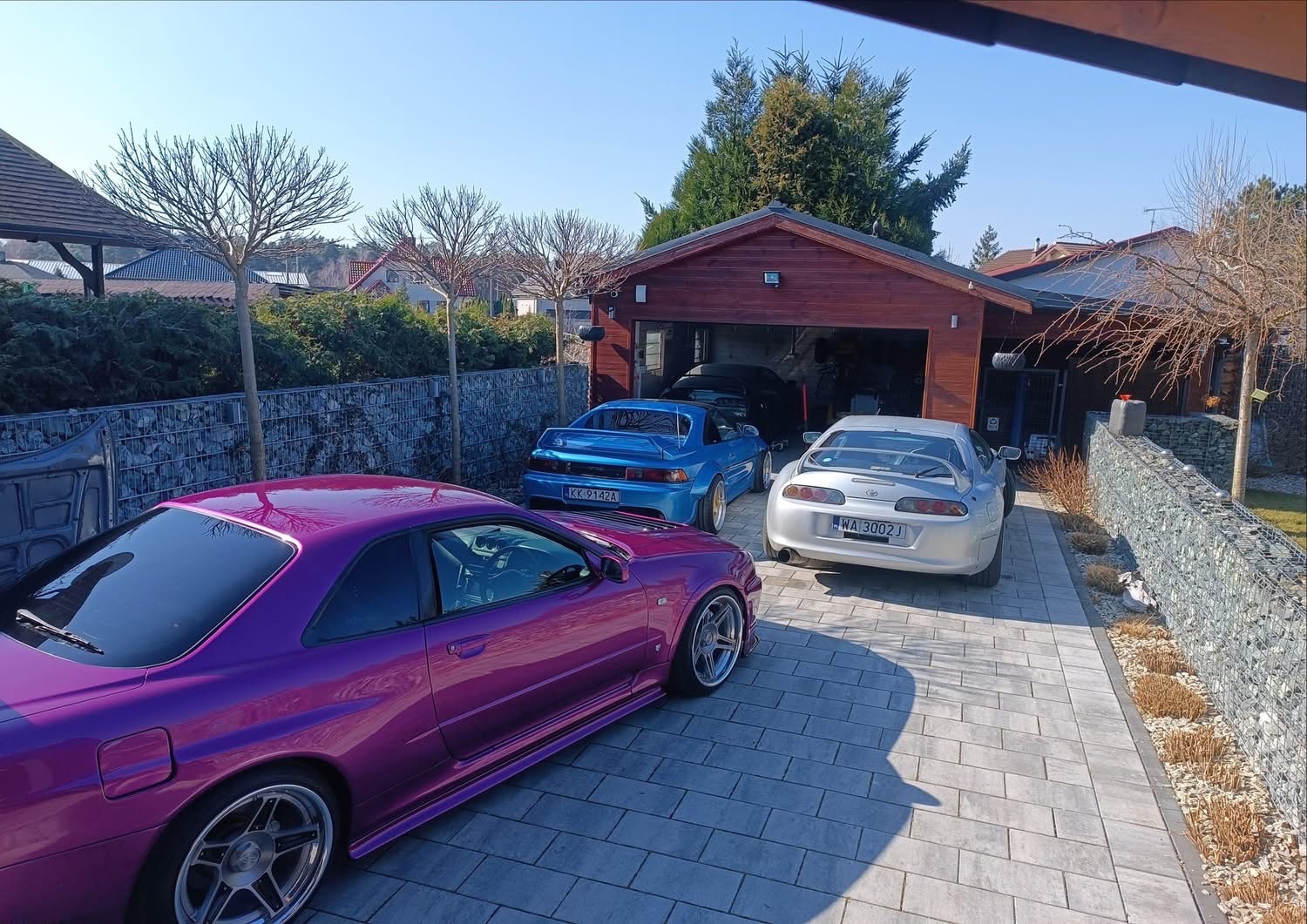 Trzy sportowe auta (fioletowy Nissan Skyline R34, niebieski Toyota MR2, srebrna Toyota Supra) zaparkowane przed garażem w słoneczny dzień, widok z boku.