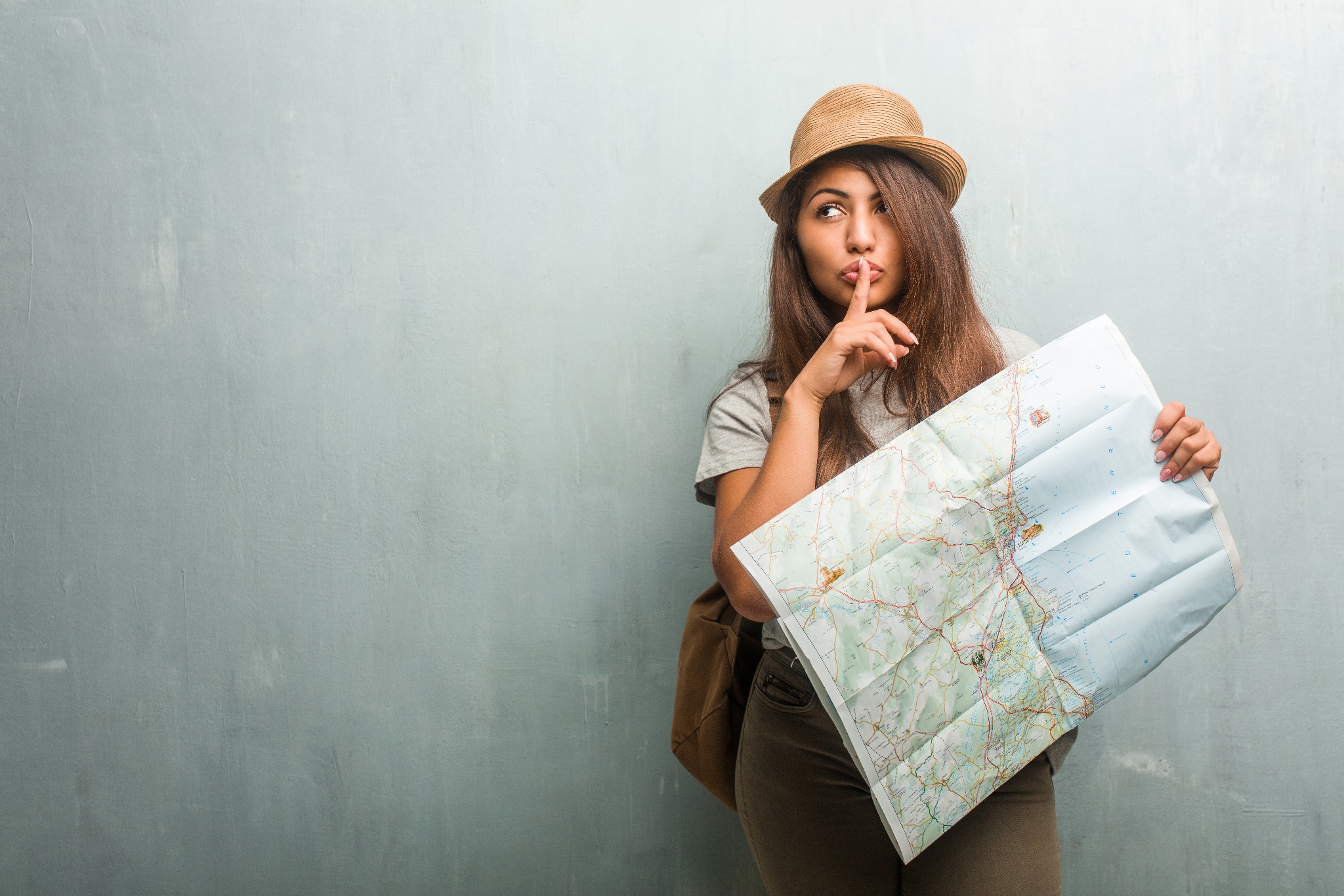 Kobieta w słomkowym kapeluszu, z plecakiem i mapą w rękach, gestem uciszenia prosi o ciszę, stojąc na tle szarej ściany.