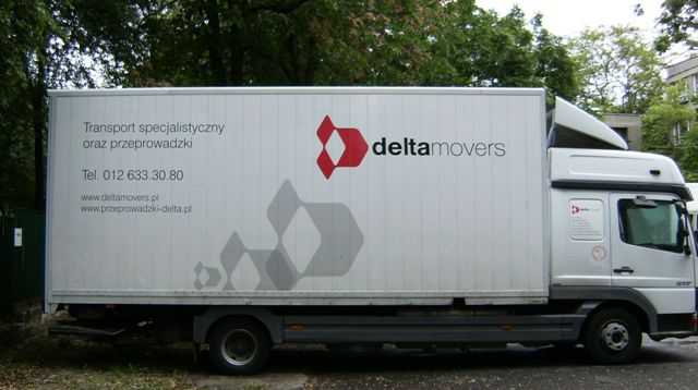 Biała ciężarówka firmy Delta Movers z czerwonym logo i informacjami kontaktowymi zaparkowana na tle zieleni miejskiej.
