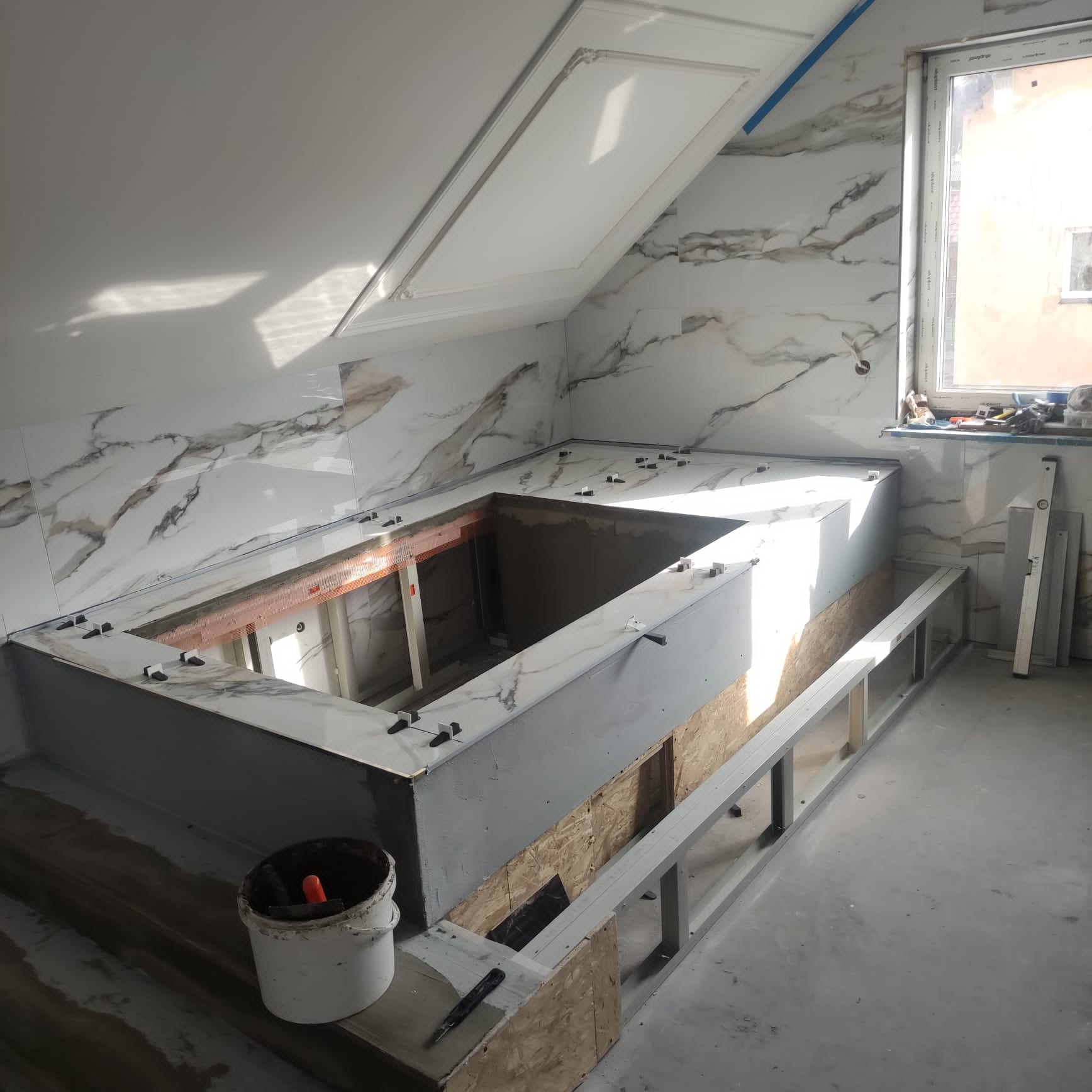 Wnętrze łazienki na poddaszu w trakcie tworzenia obudowy wanny z marmurowych płytek, z widocznym stelażem metalowym, wiadrem z zaprawą i poziomnicą przy oknie.