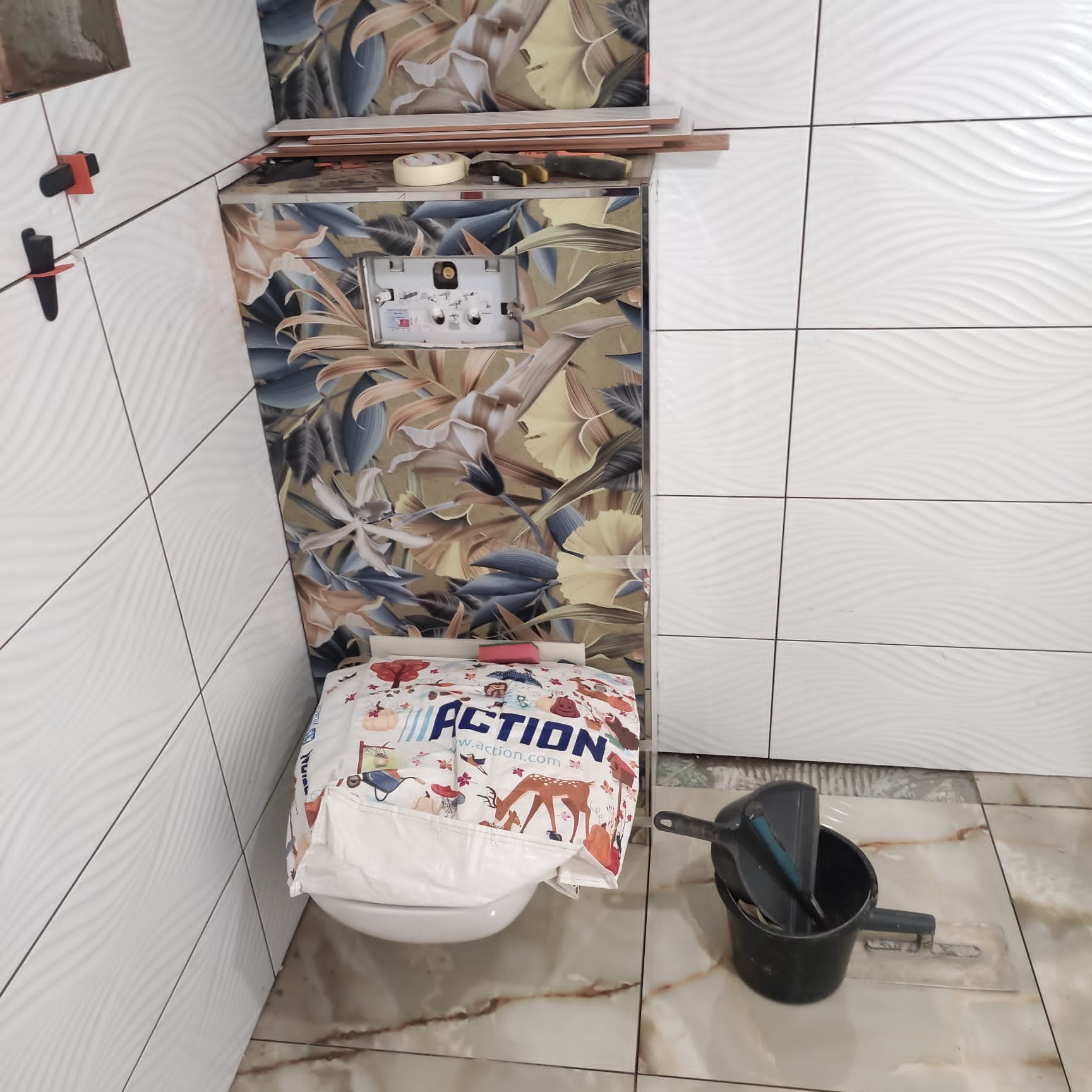 Montaż stelaża podtynkowego WC z wzorem kwiatowym, częściowo wykończone białe płytki z falistą strukturą, narzędzia na stelażu, wiadro i kielnia na podłodze z marmurowym wzorem, zabezpieczona toaleta.