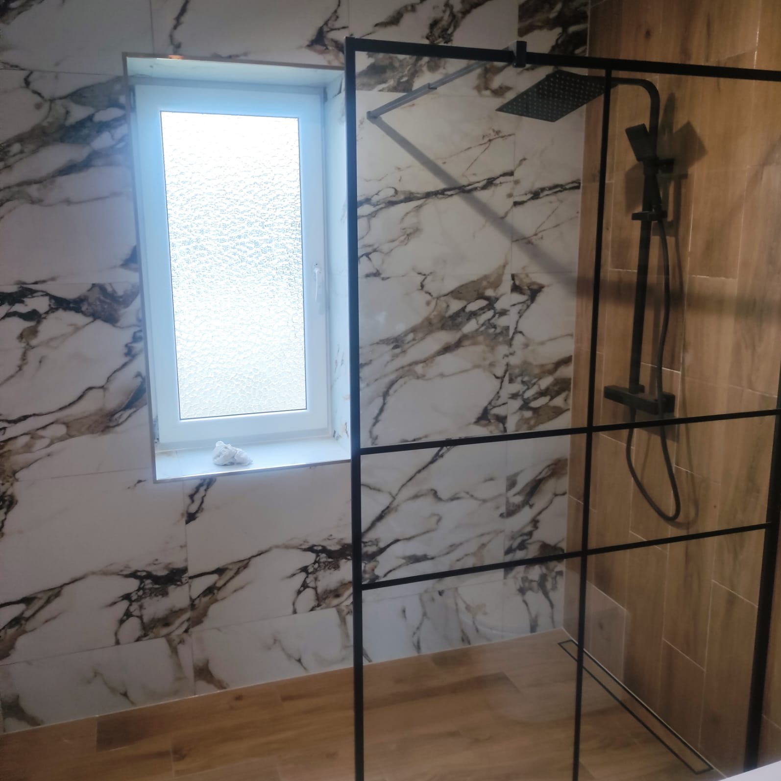 Nowoczesna kabina prysznicowa z czarną armaturą i ramą, marmurowe płytki na ścianach, okno z matową szybą i drewnopodobna podłoga.