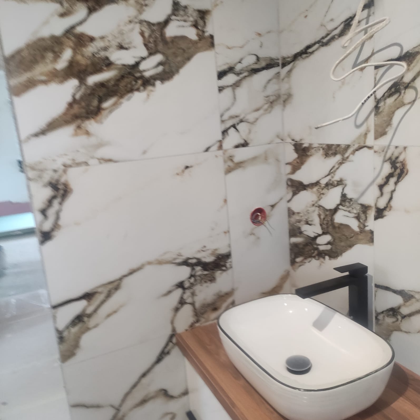 Umywalka nablatowa w kształcie zaokrąglonego prostokąta z czarną armaturą na drewnianym blacie, na tle marmurowych płytek ściennych z brązowymi żyłami, widoczne wystające przewody elektryczne.