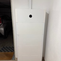 Mateusz Puchalski - Biały, modułowy magazyn energii w garażu, widoczny samochód i dywanik przy drzwiach.