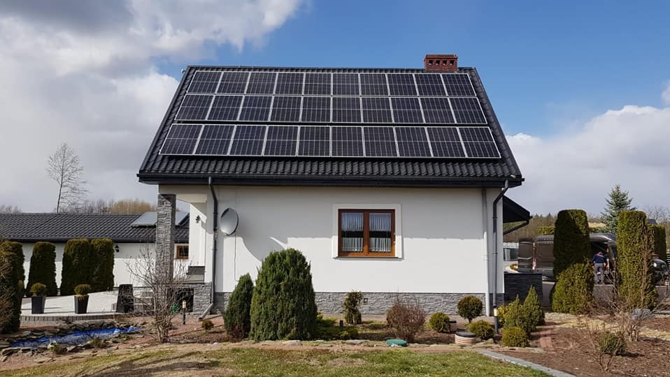 Dom jednorodzinny z panelami słonecznymi na dachu, widok z ogrodu w słoneczny dzień, z widoczną anteną satelitarną na ścianie.