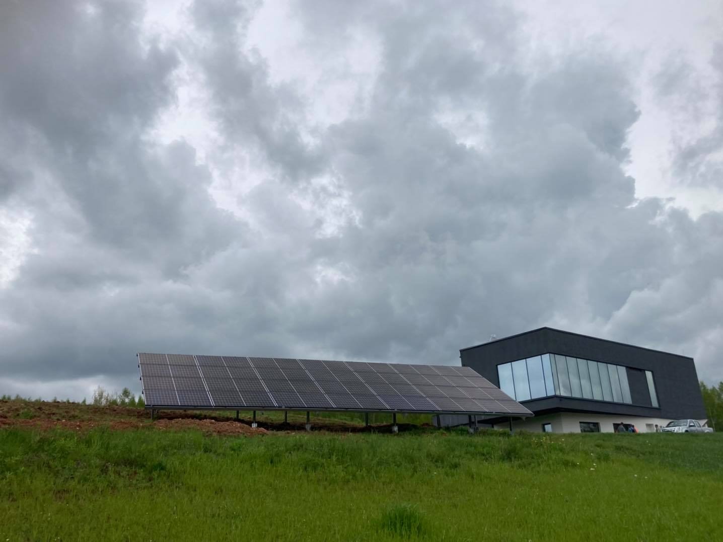 Panel słoneczny na metalowej konstrukcji, ustawiony na wzgórzu z trawą, obok nowoczesnego, ciemnego budynku z dużymi oknami, pod pochmurnym niebem.