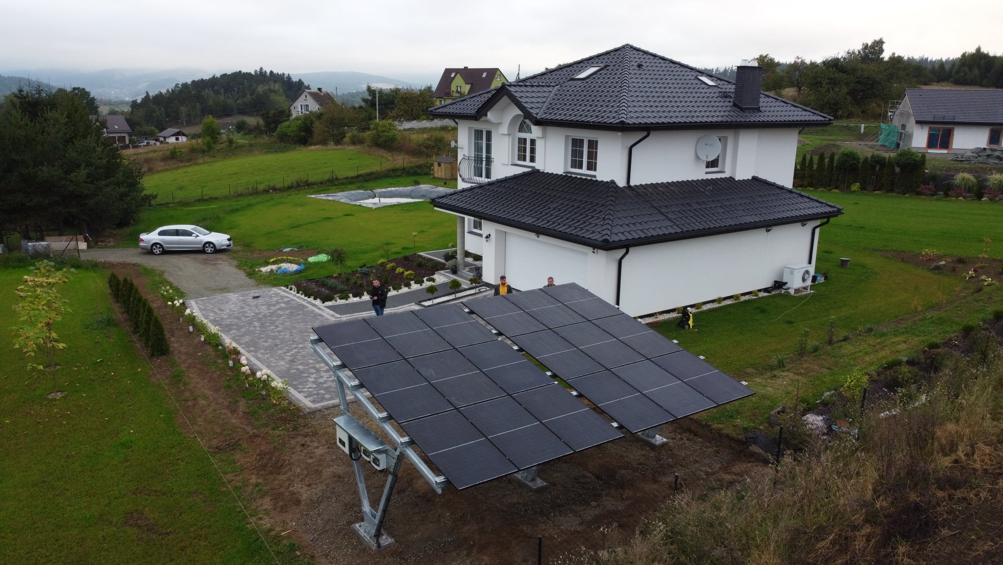 Instalacja fotowoltaiczna na gruncie z widokiem na nowoczesny dom jednorodzinny i górzysty krajobraz w tle.