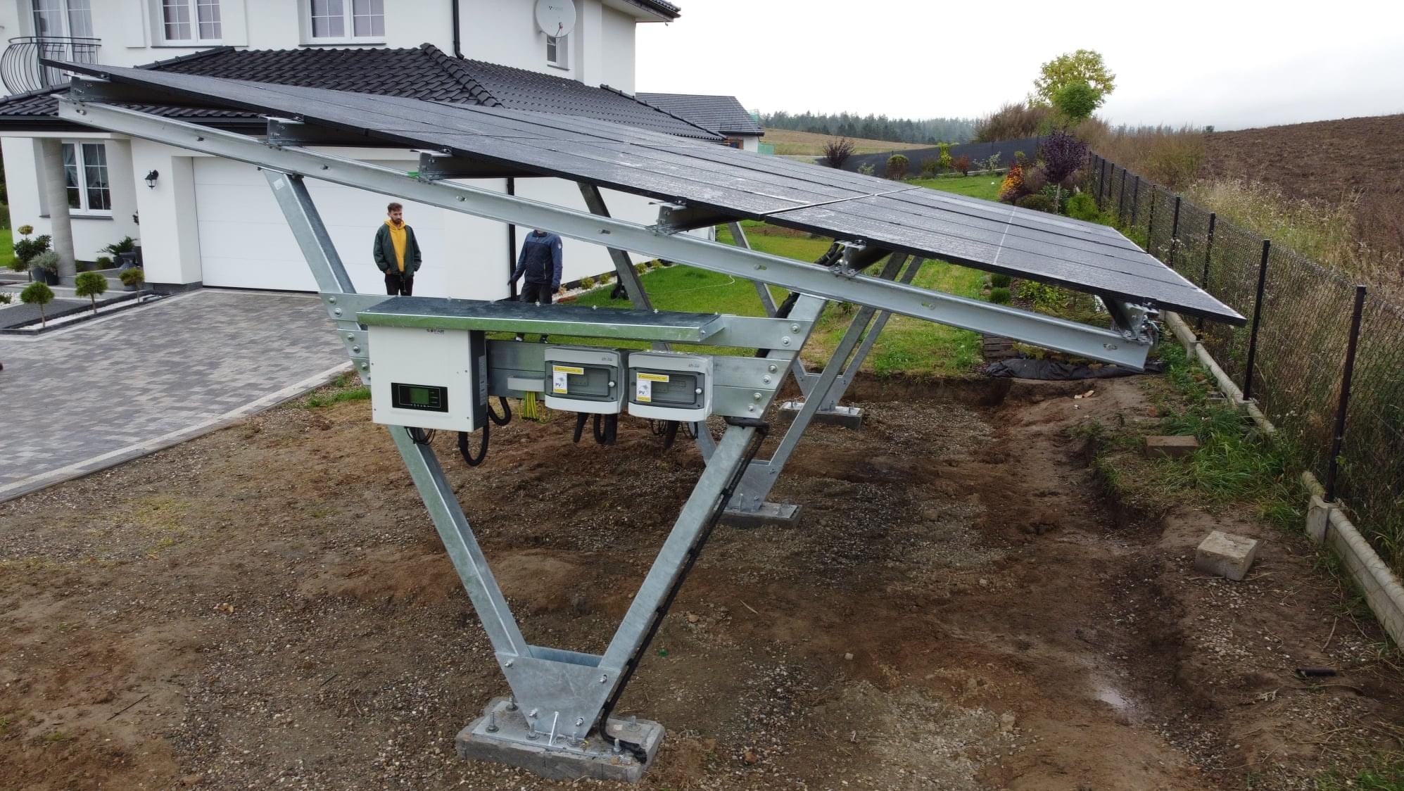 Montaż paneli słonecznych na metalowej konstrukcji w ogrodzie, widoczni dwaj pracownicy w tle, teren wokół rozkopany, instalacja elektryczna z puszkami przy panelach.