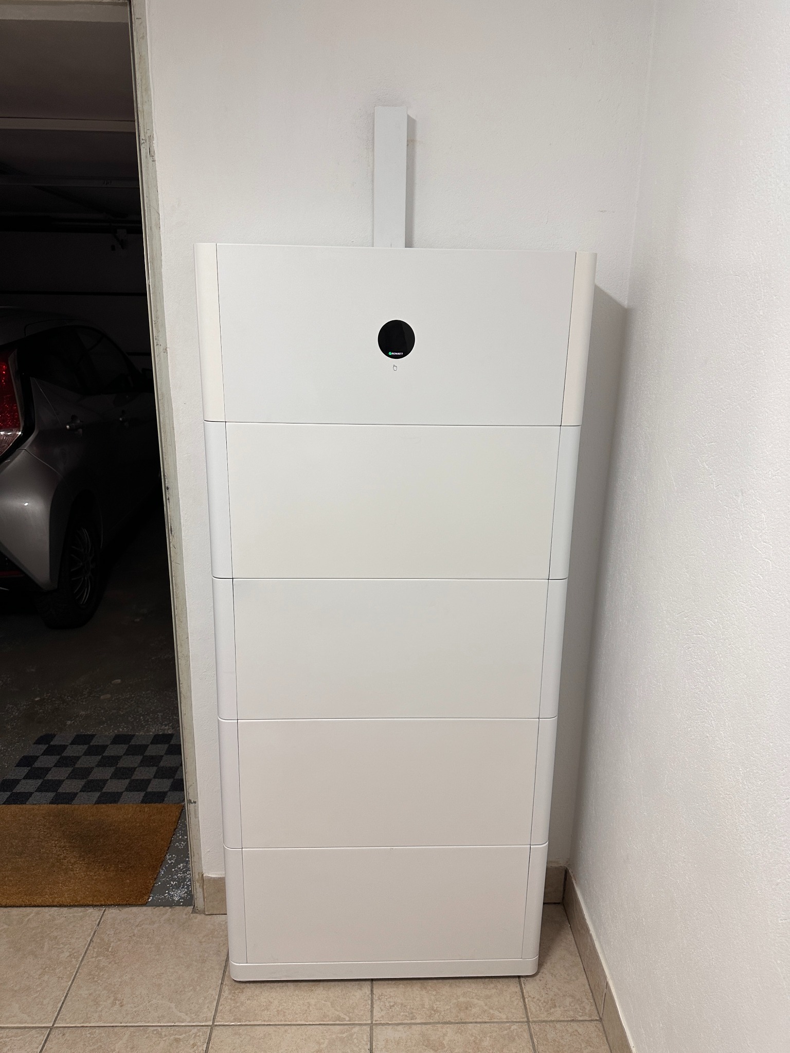 Biały, modułowy magazyn energii w garażu, widoczny samochód i dywanik przy drzwiach.