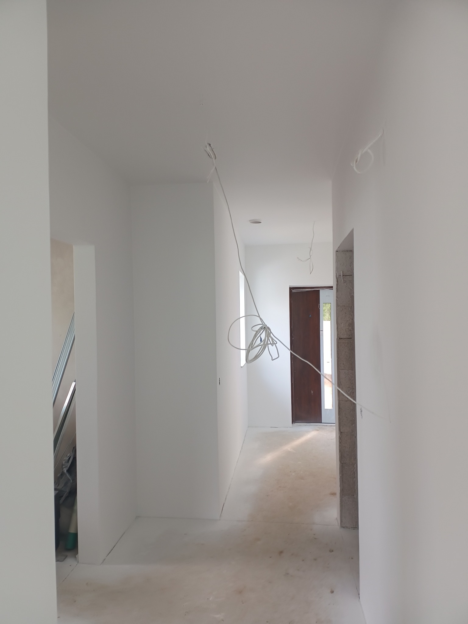 Wnętrze korytarza w trakcie prac wykończeniowych, widoczne świeżo pomalowane ściany na biało, wystające kable elektryczne i surowa podłoga, zarys futryny drzwiowej bez ościeżnicy.