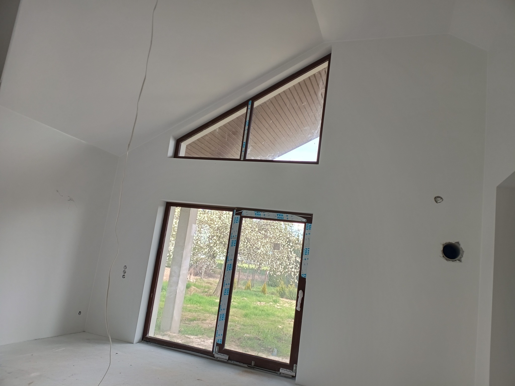 Wnętrze świeżo wykończonego pokoju z białymi ścianami, brązowymi ramami okiennymi, widokiem na kwitnące drzewo przez duże okno przesuwne i trójkątne okno pod sufitem.