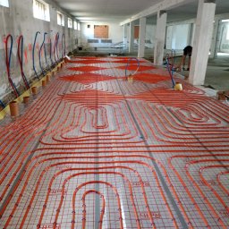 INNS SPÓŁKA Z OGRANICZONĄ ODPOWIEDZIALNOŚCIĄ - Instalacja rur grzewczych w systemie podłogowym na folii z siatką, widoczne rury doprowadzające w kolorach czerwonym i niebieskim, surowy stan budynku.