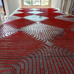 Czerwone rurki systemu rozprowadzania ciepła ułożone spiralnie na srebrnej folii izolacyjnej, widoczne okno w tle, instalacja w surowym stanie.